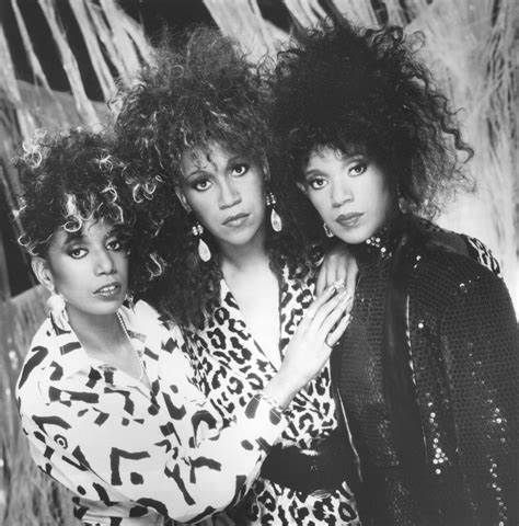 Three women posing together in patterned outfits, with voluminous curly hair and serious expressions.