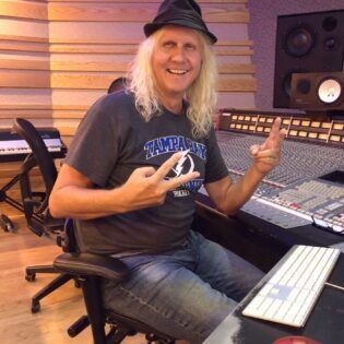 Person smiling in a recording studio, seated at a mixing console and gesturing with both hands.