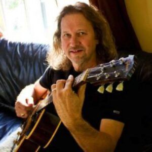 Man playing acoustic guitar on a couch, smiling indoors with warm light.