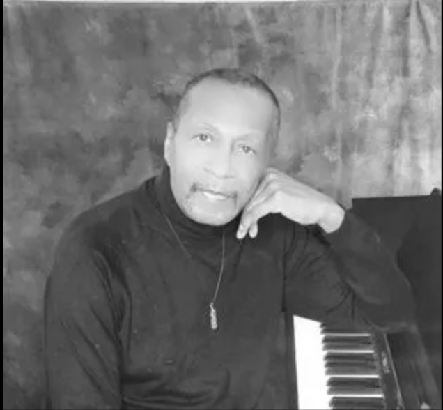 Man seated beside a piano, resting his head on his hand in a black turtleneck against a textured backdrop
