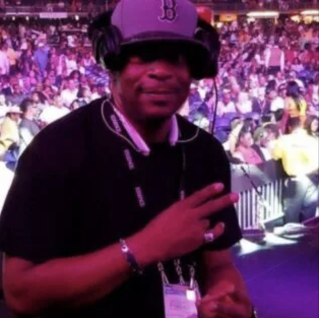 Person in a black jacket and cap making a peace sign in a crowded arena with purple lighting