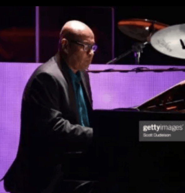 Pianist seated at a grand piano on a purple-lit stage, performing in profile