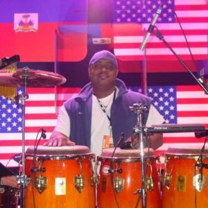 Drummer playing orange conga drums on stage with U.S. flag backdrop and purple lighting