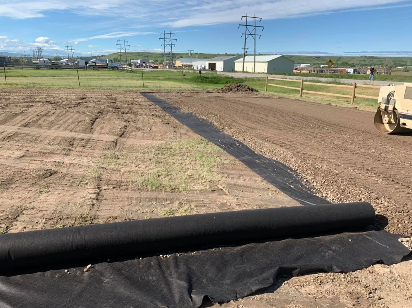 A roll of black landscape fabric lies on dirt at a construction site, with a roller vehicle visible in the background.