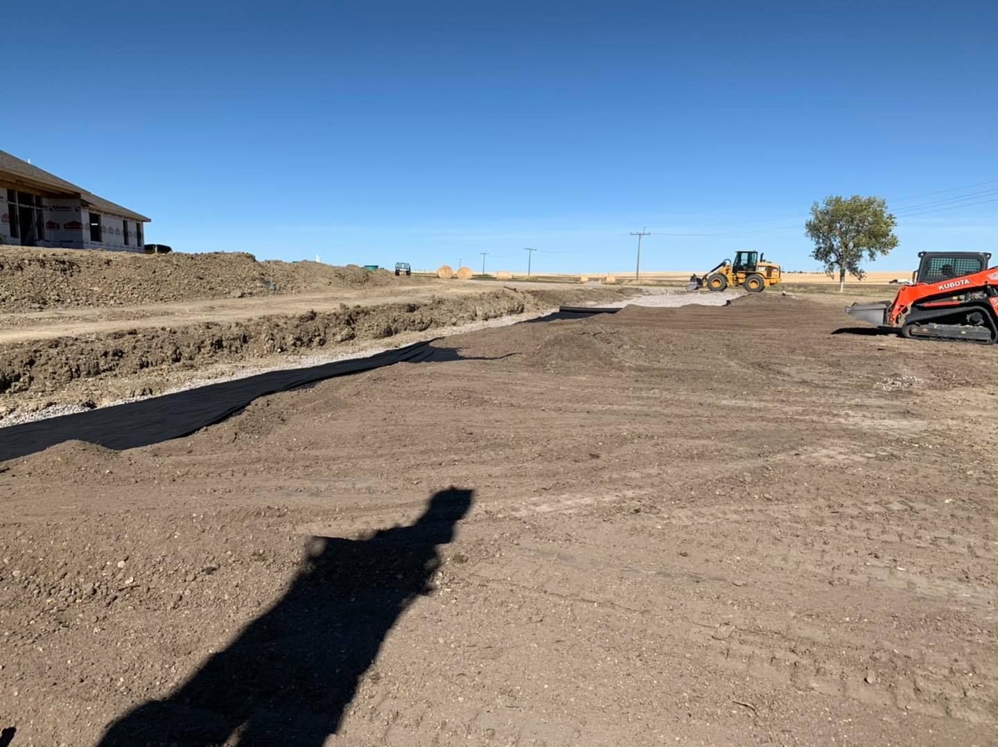 Construction site with a dirt lot, heavy equipment, and a partially built house under a clear blue sky.