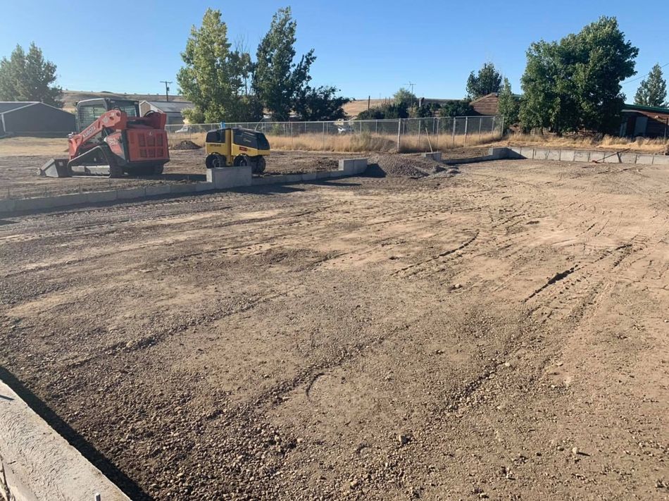 A construction site with a concrete foundation, a red skid steer, and a yellow plate compactor under a clear blue sky.