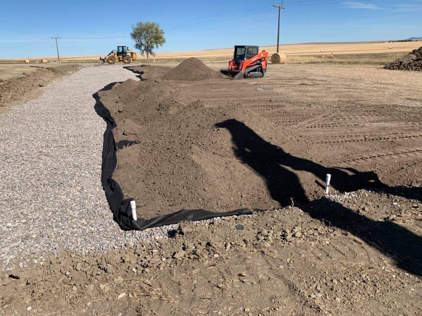 Construction site with a gravel path, black erosion control fabric, and construction equipment under a clear blue sky.