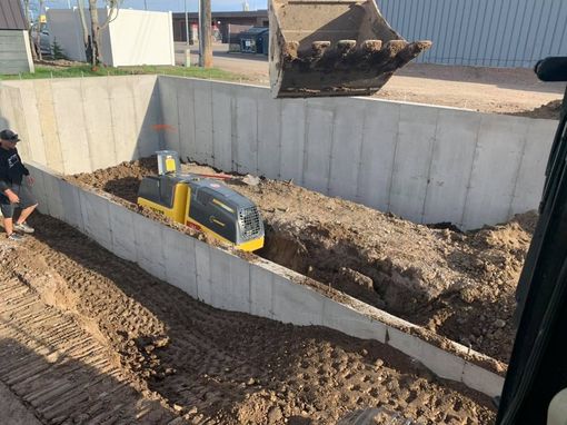 An excavator bucket hovers above a construction site with a yellow mini-excavator working inside a concrete foundation.