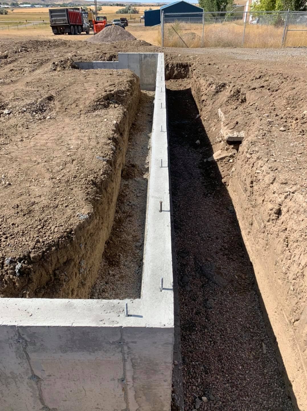 A concrete foundation wall sits between two deep trenches in an outdoor construction site with a truck in the distance.