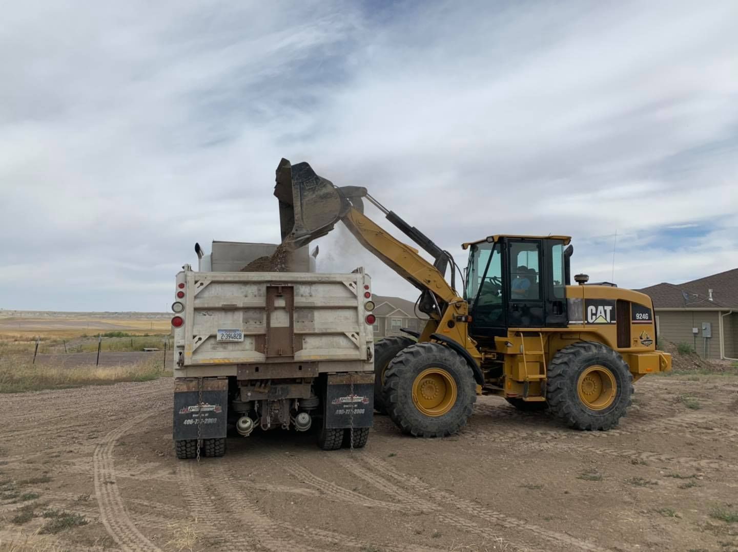 A yellow CAT front-end loader dumps dirt into the back of a parked dump truck on a dirt lot under a cloudy sky.