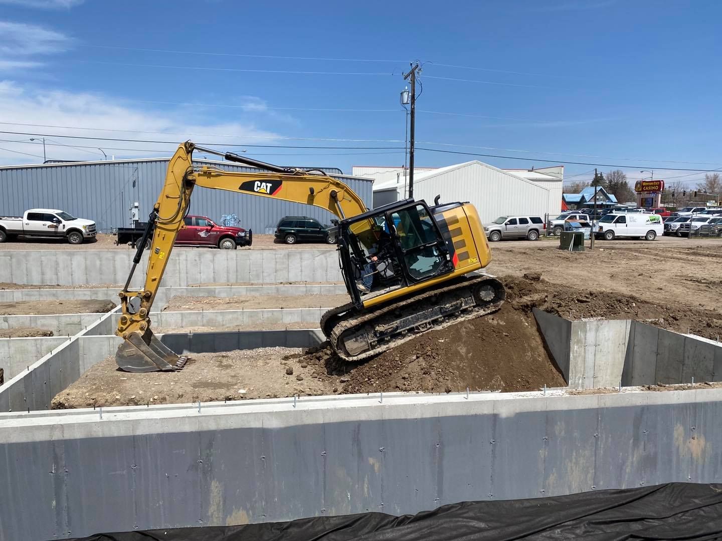 A yellow Caterpillar excavator sits on a mound of dirt inside a concrete foundation at a construction site.