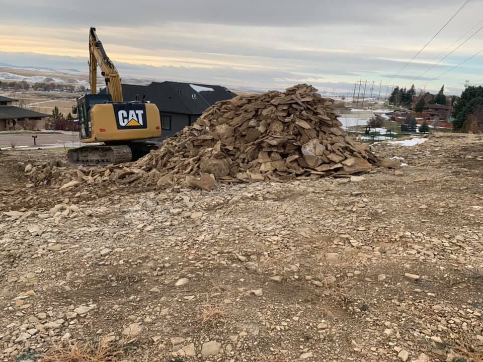 A yellow Caterpillar excavator sits on a rocky, uneven construction site next to a large pile of excavated stone.