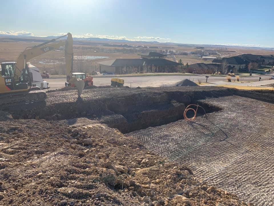 A yellow excavator digs a large trench at a residential construction site on a sunny day.
