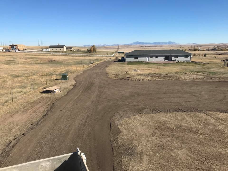 A dirt driveway construction area in a dry, open rural landscape with houses and mountains in the distance.