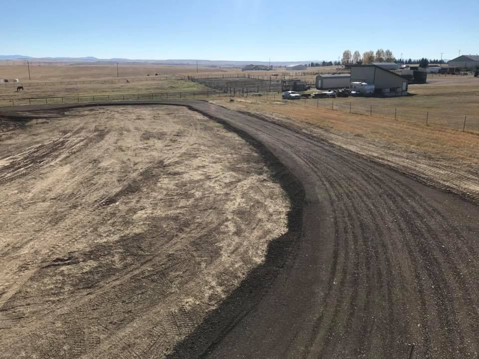 A freshly graded dirt road curves through a dry, rural field toward distant buildings under a clear blue sky.