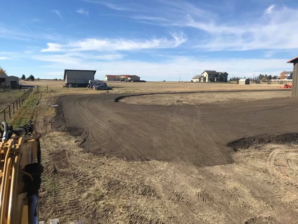 Construction equipment parked in a dry, open field with a freshly graded dirt road curve under a bright blue sky.
