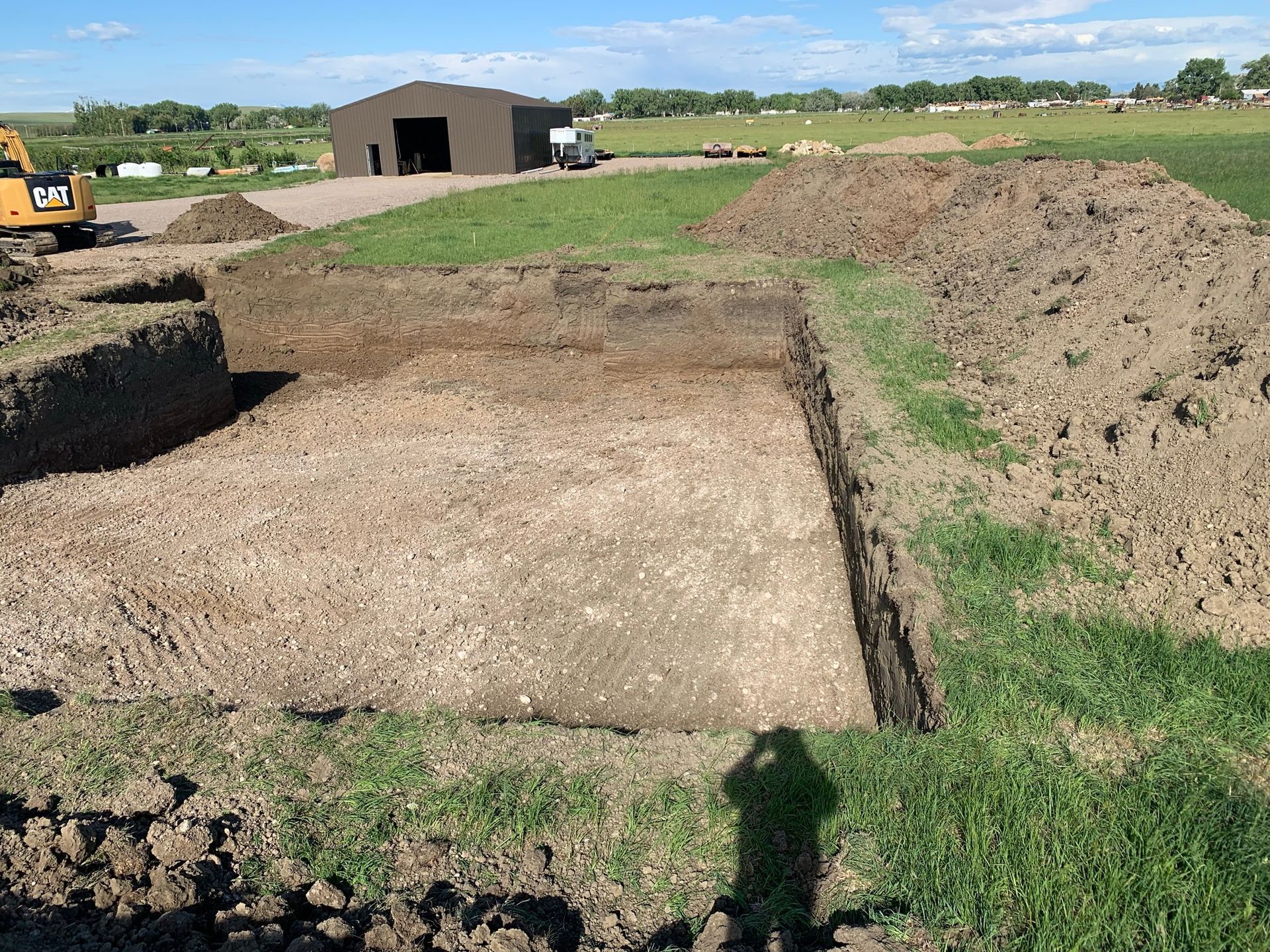 An open rectangular excavation pit in a grassy field, with dirt piles and a large metal shed in the background.