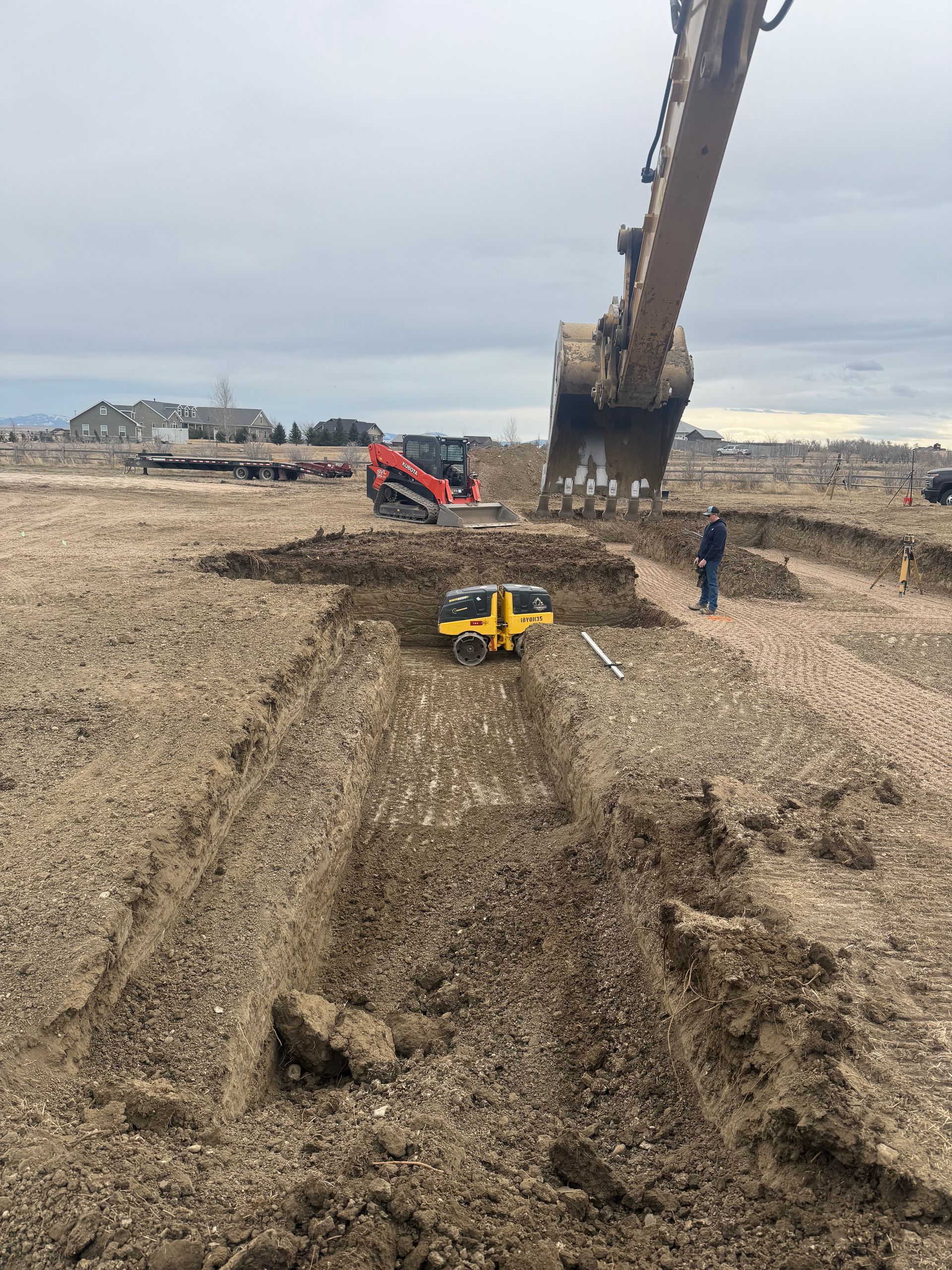 A large excavator, a red skid steer, and a yellow compactor working on a dirt construction site under a cloudy sky.