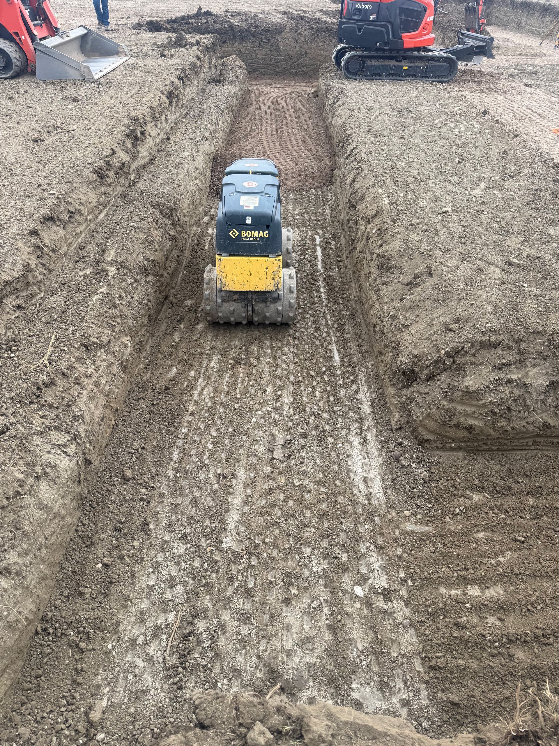 A yellow and black skid-steer loader driving through a dirt trench at a construction site with other machinery nearby.
