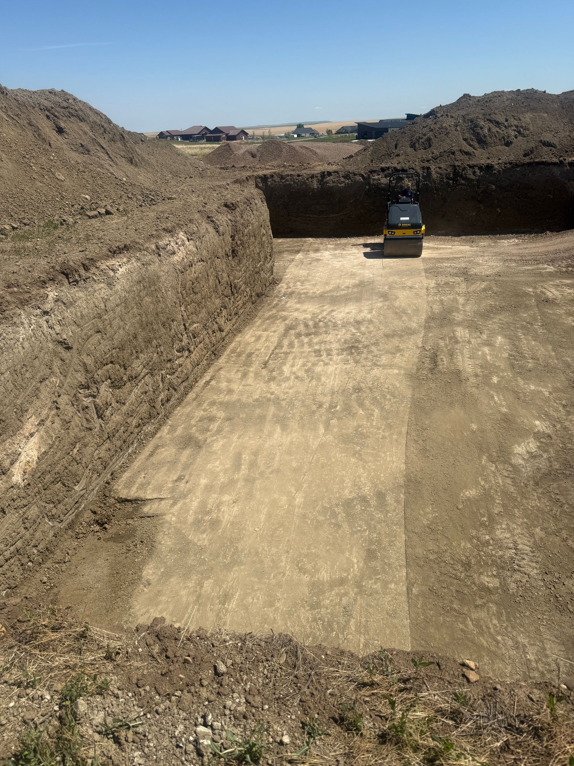 A construction excavator sits in a large, excavated rectangular dirt pit on a sunny day.
