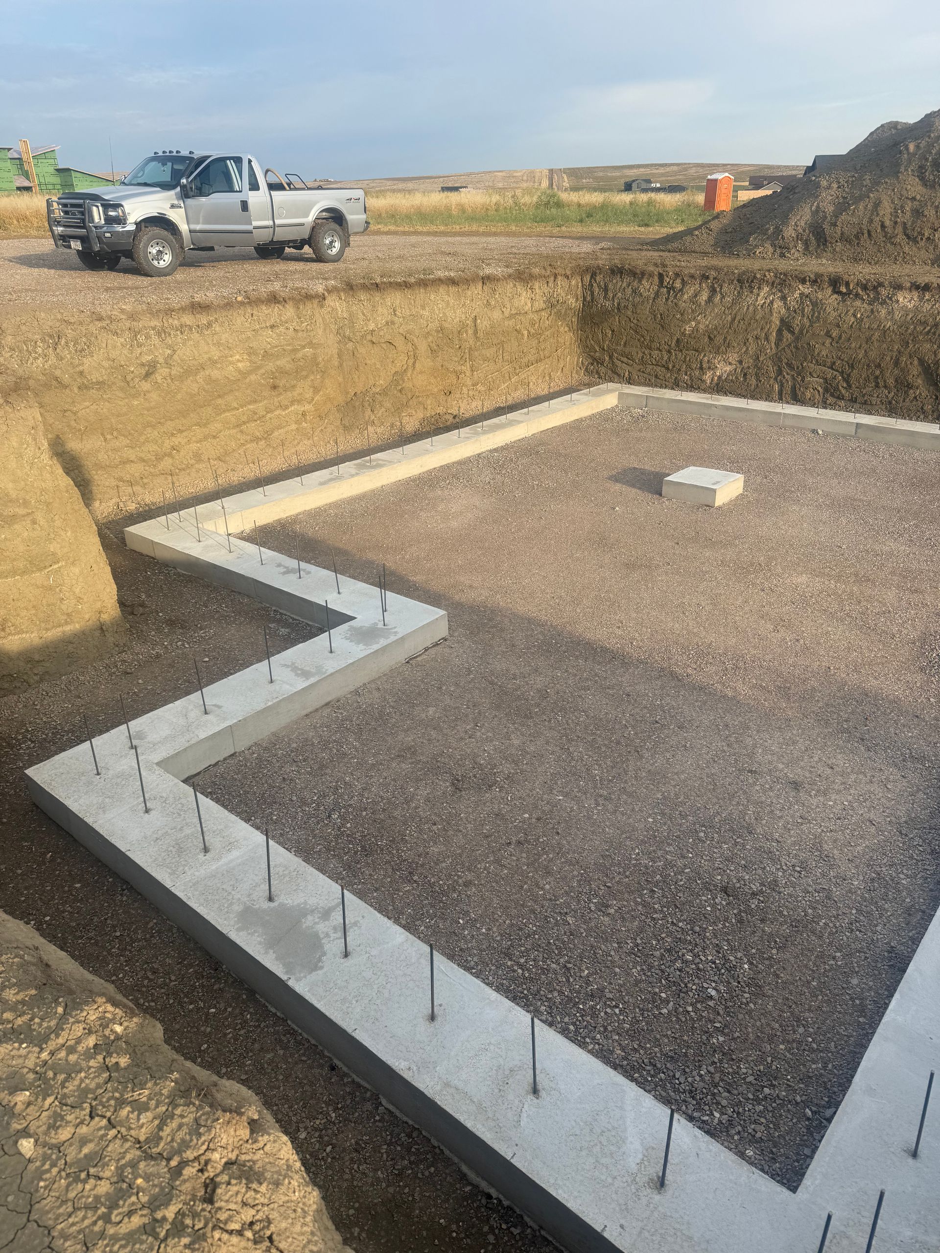 A concrete foundation for a building with metal rebar studs, set in a dirt excavation site with a pickup truck nearby.
