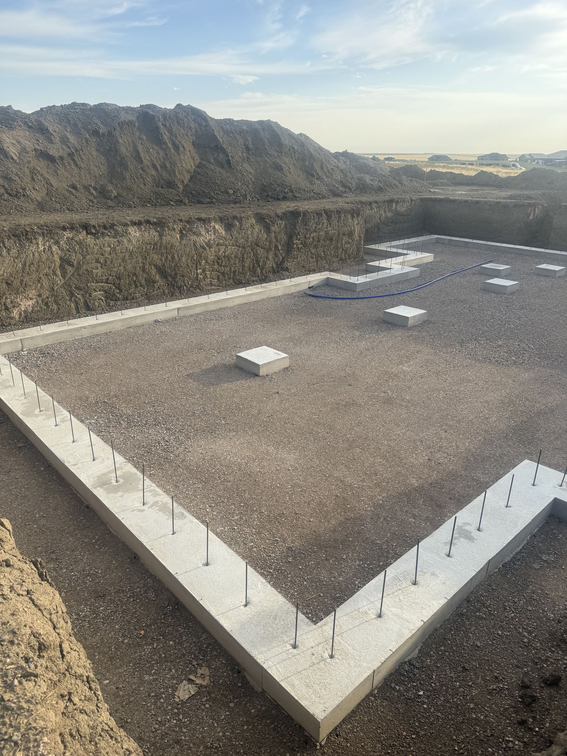A concrete foundation for a building under construction, featuring anchor bolts and gravel fill in a large, dirt-walled pit.