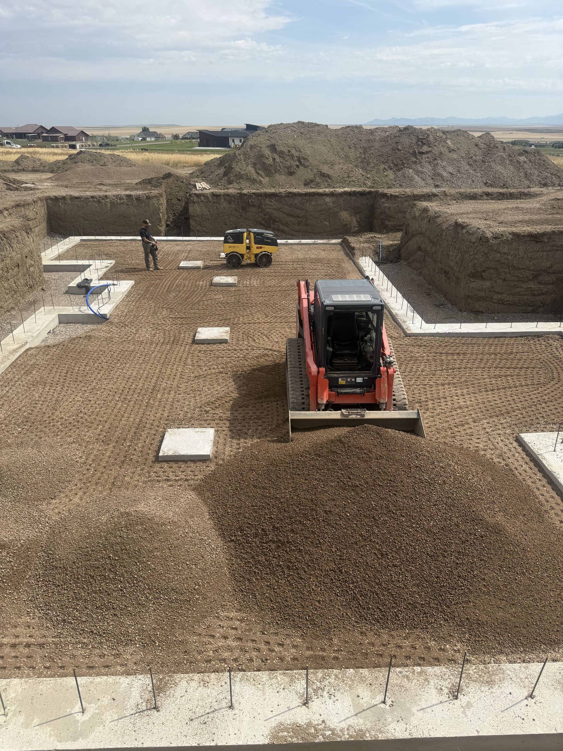 An orange skid steer levels gravel inside a building foundation under construction, with a small roller nearby.
