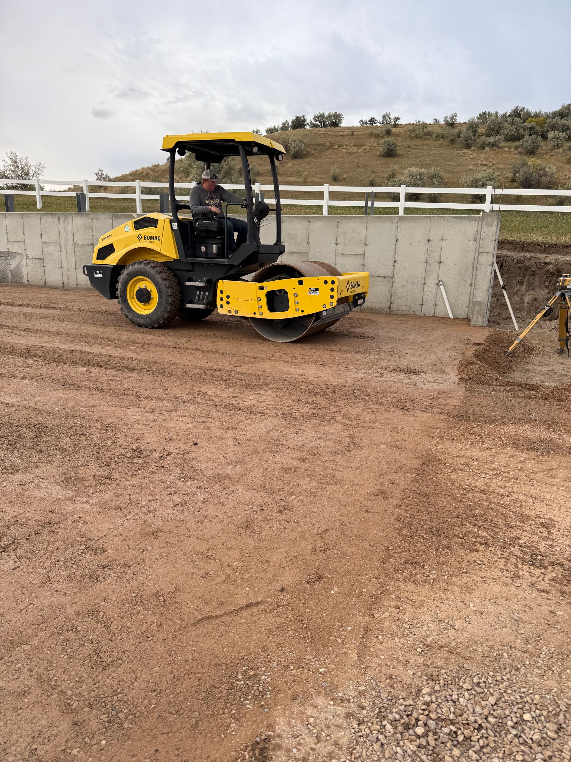 A yellow soil compactor machine drives across a dirt construction site next to a concrete retaining wall.