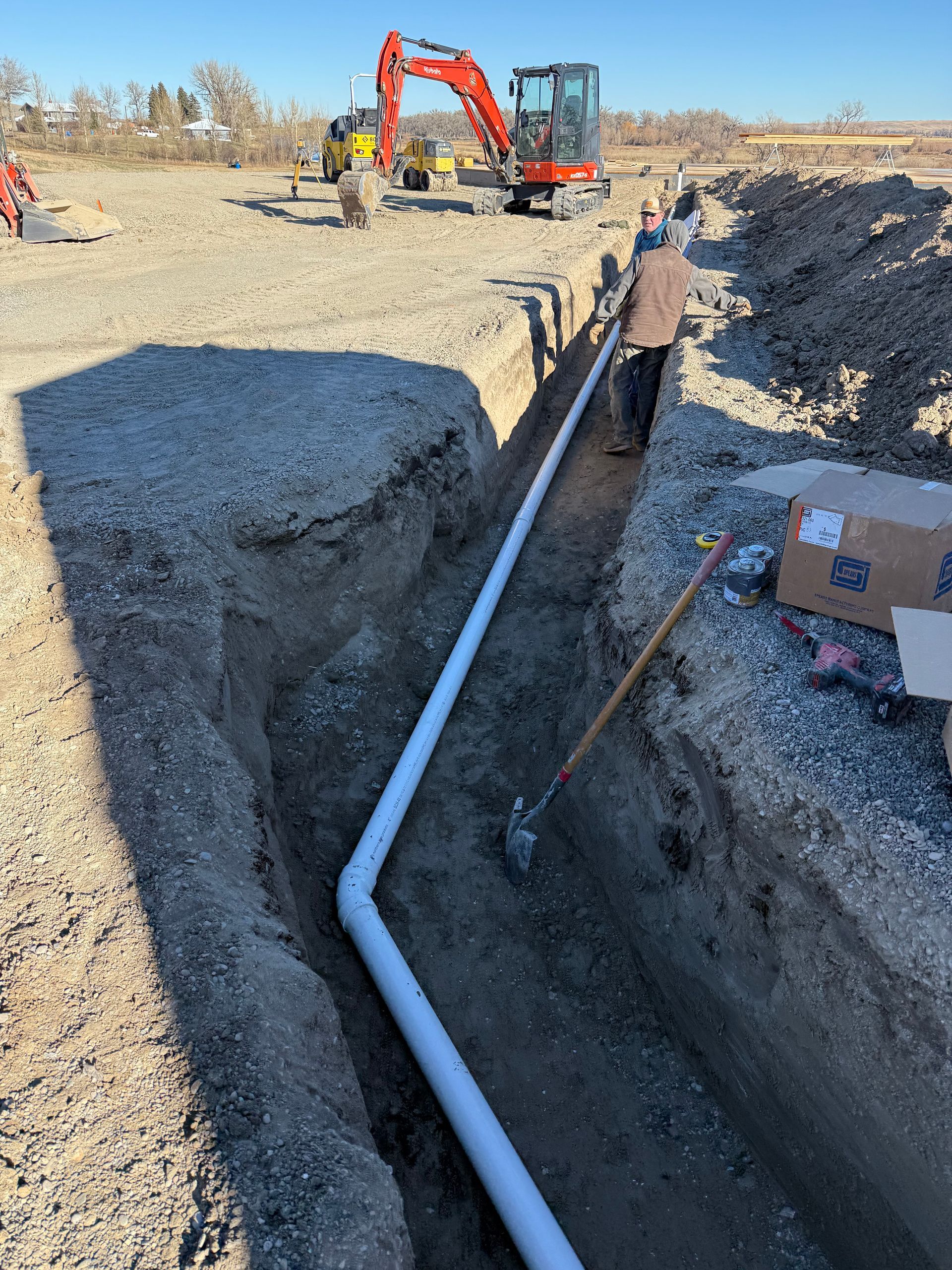 Construction worker installing white PVC pipe in a narrow dirt trench with an excavator visible in the background.