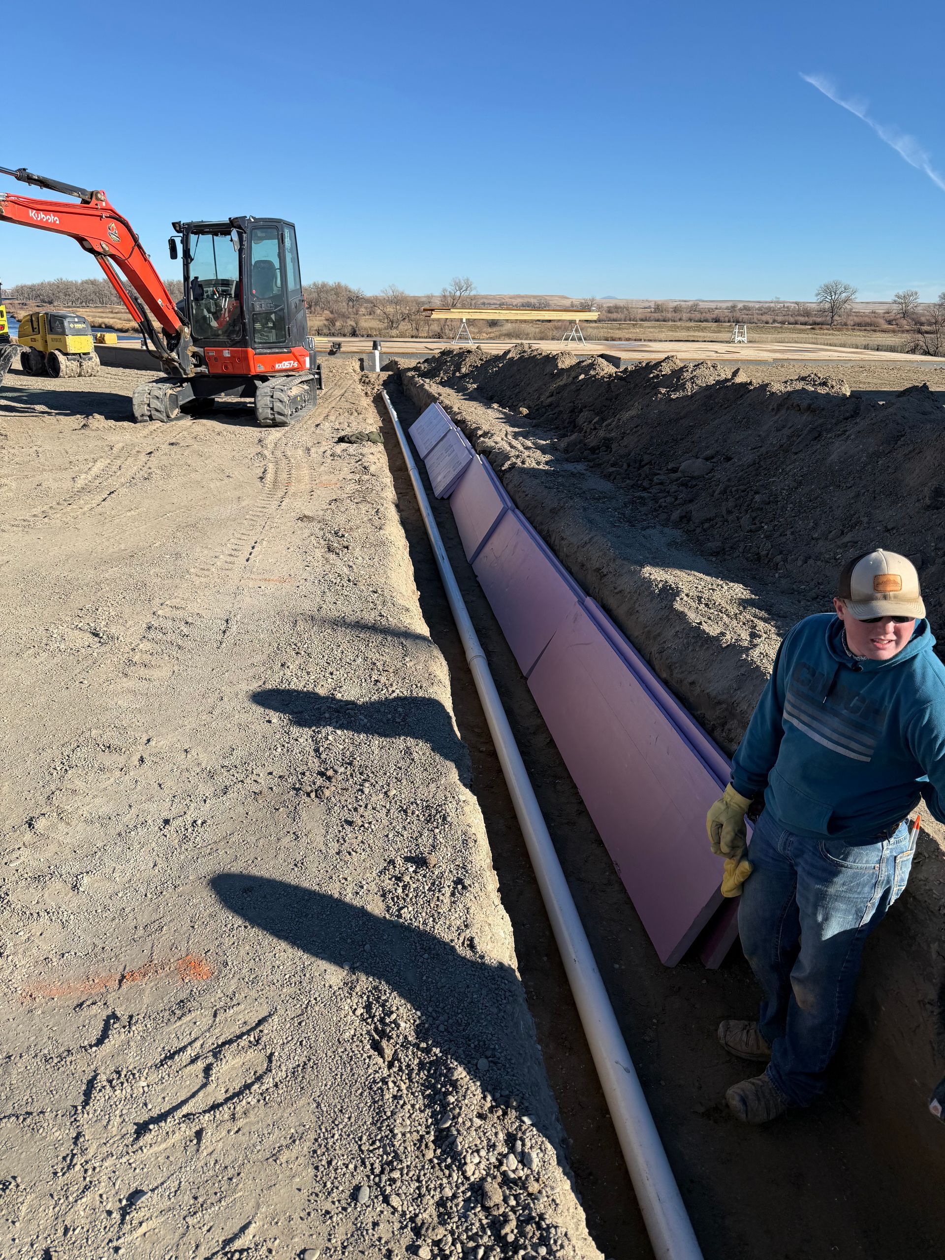 A person wearing a blue hoodie and cap stands in a trench next to a white pipe and pink insulation boards near an excavator.