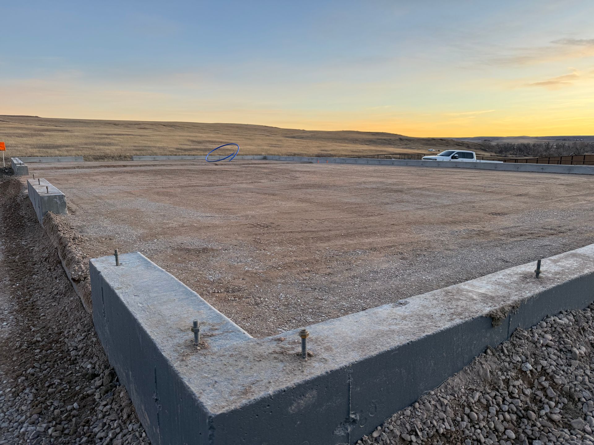 A concrete foundation for a building under construction, featuring exposed anchor bolts on a gravel-filled site at sunset.