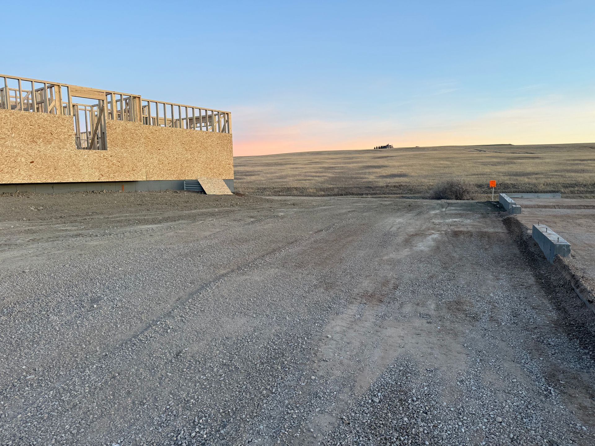 A wooden house frame under construction sits on a gravel lot at sunrise, with a vast, open field in the distance.