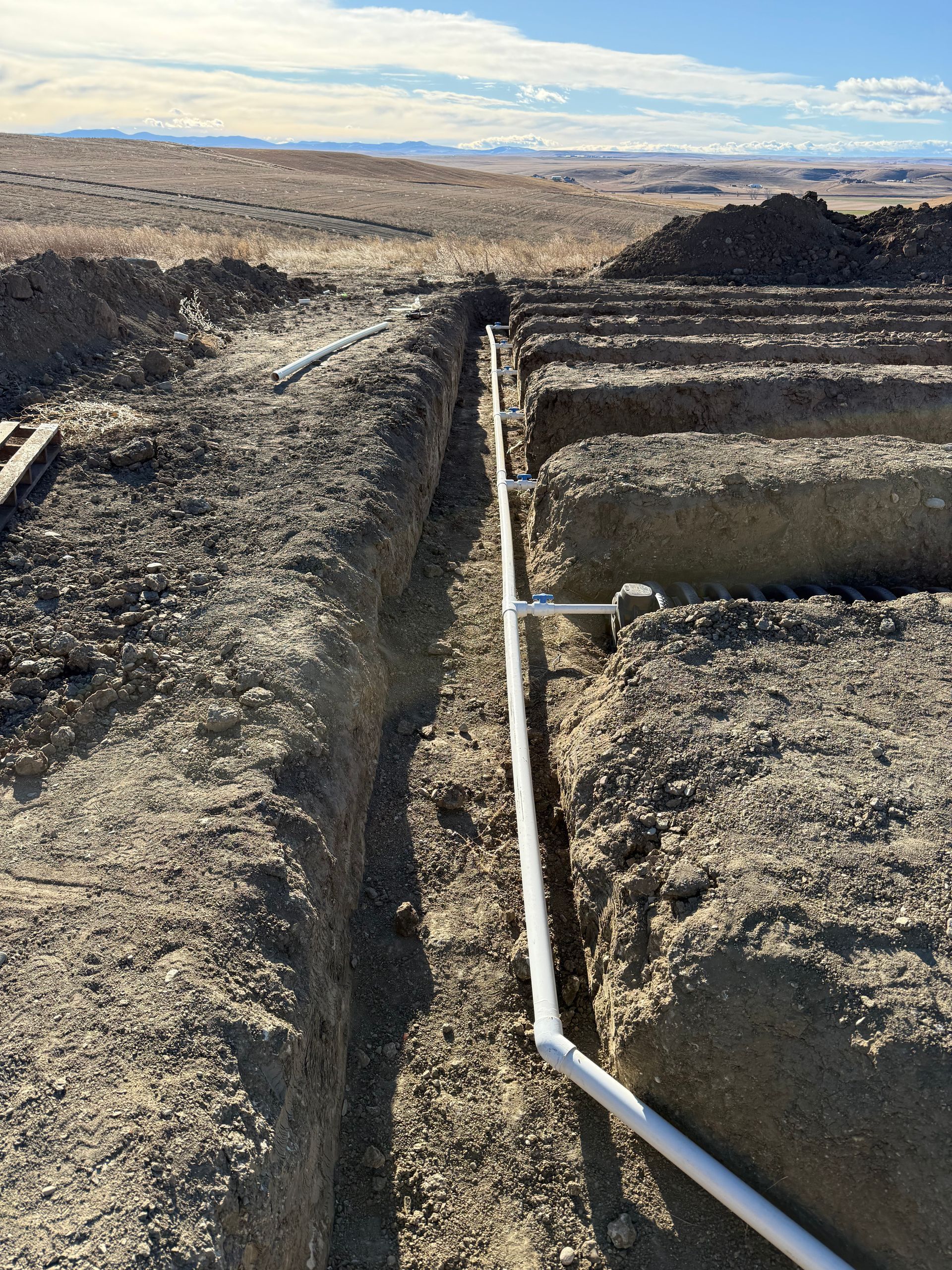 White PVC pipes lie in a trench alongside raised dirt rows at a construction or irrigation site under a blue sky.