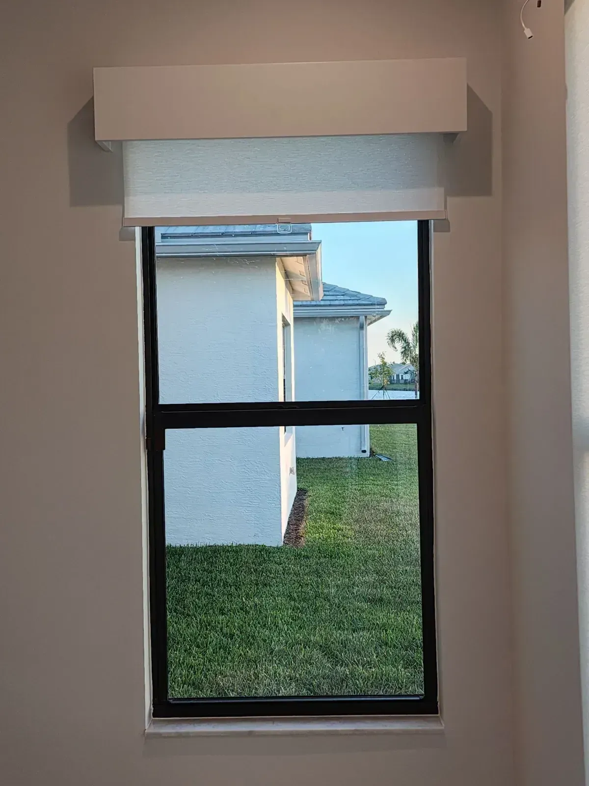 Window with a partially lowered white blind overlooking a grassy yard and neighboring house.