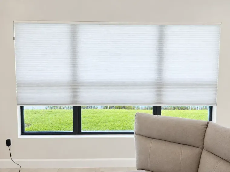 White pleated blinds over a window with green grass outside and a beige sofa in the corner