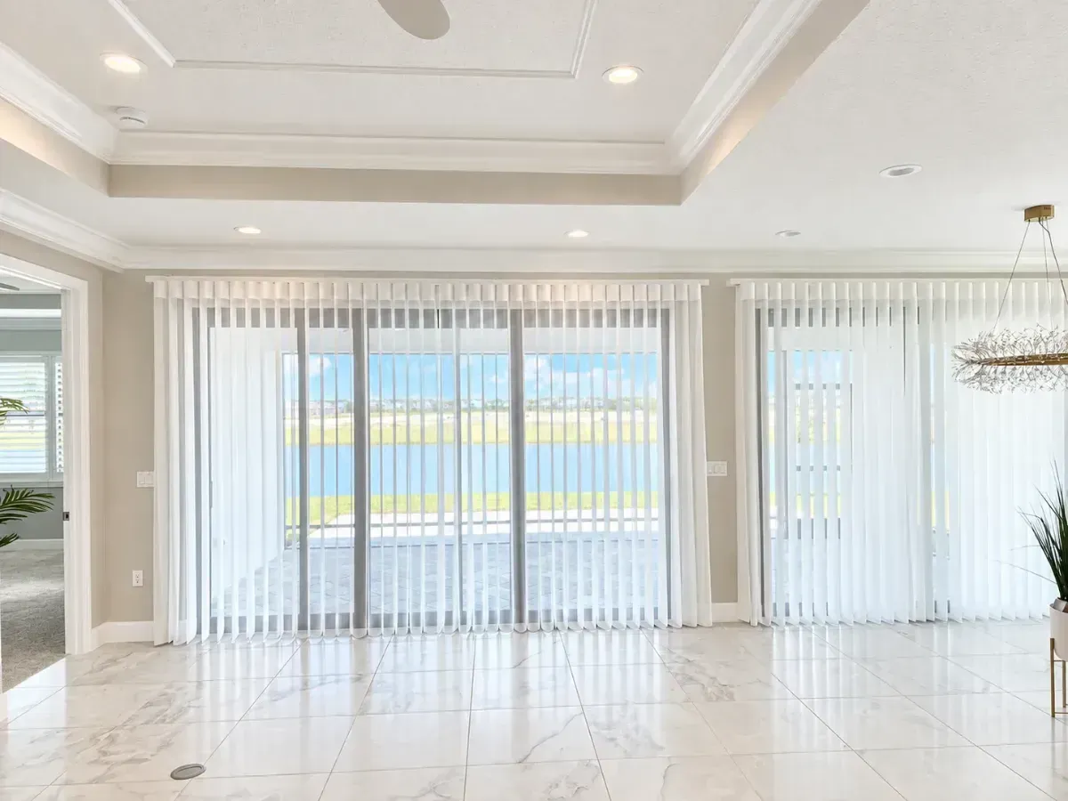 Bright empty living room with large sliding glass doors and vertical blinds, light tile floor, and tray ceiling