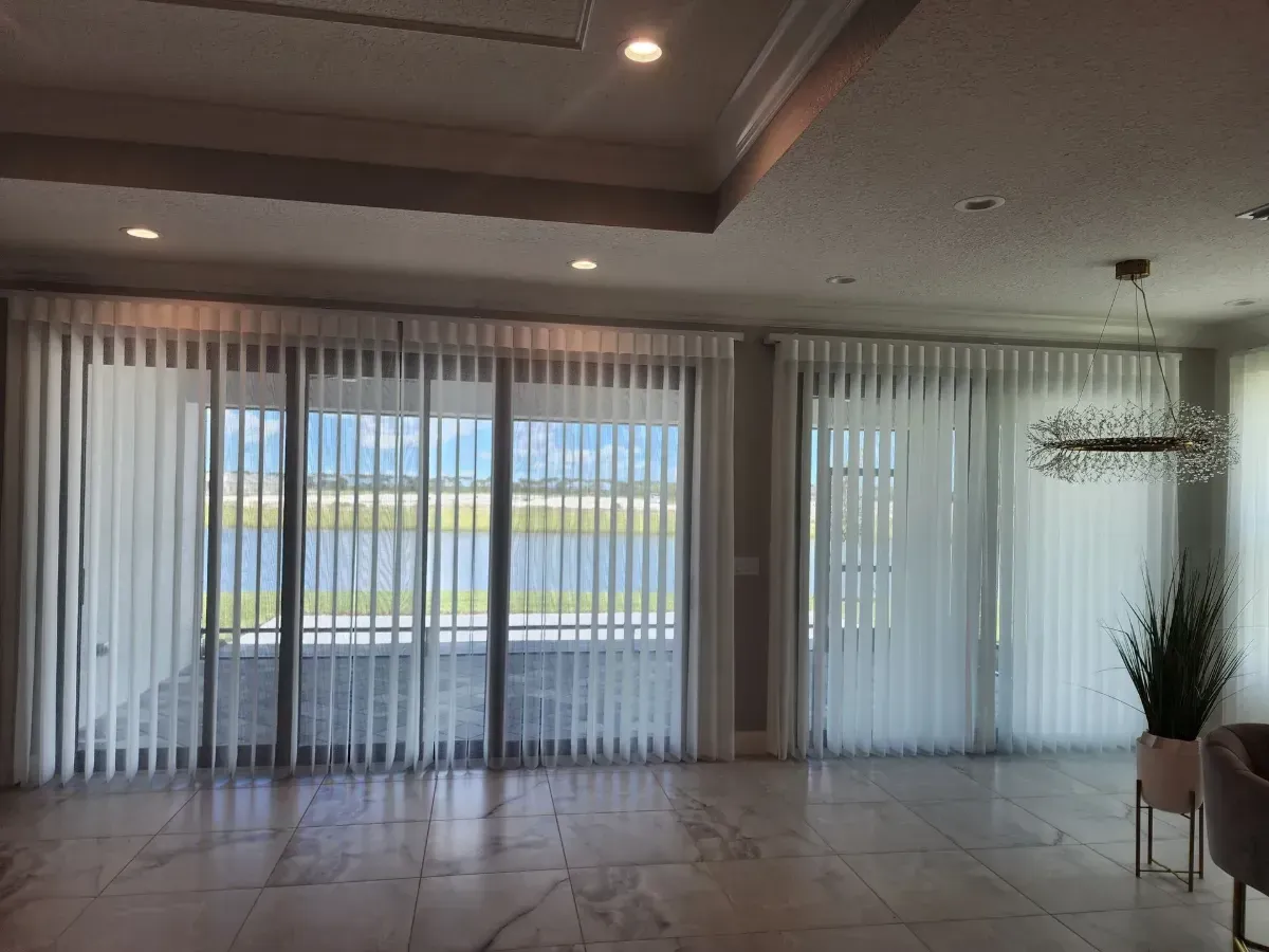 Spacious empty living room with tiled floor, sheer curtains, recessed ceiling lights, and a chandelier.