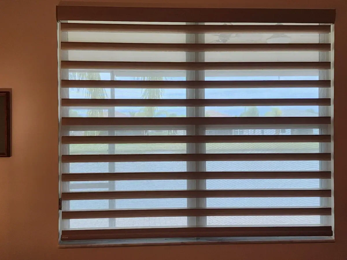 Window with horizontal blinds partially open, letting in daylight against a beige wall.