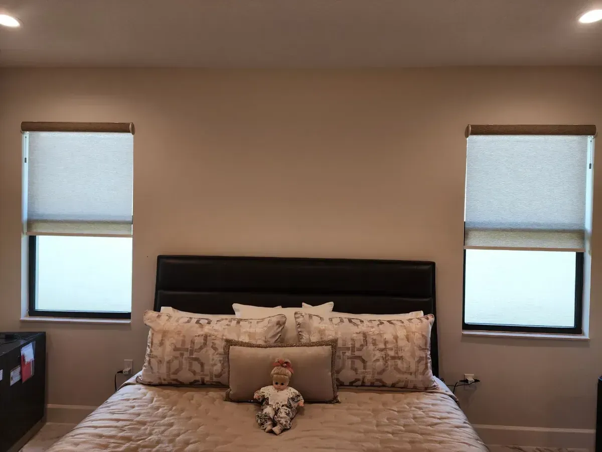 Bedroom with a black headboard, beige bedding, and two windows with closed blinds.