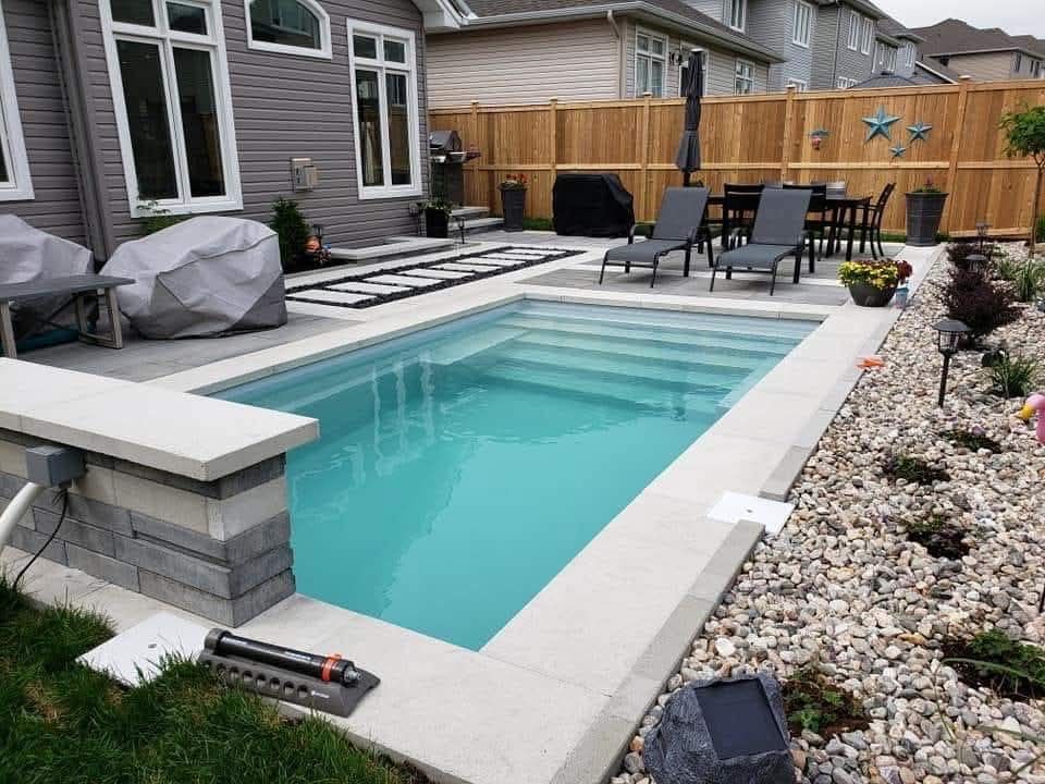 Un jardin avec une piscine rectangulaire creusée, des margelles en pierre gris clair, du mobilier de terrasse et un jardin de rocaille décoratif.