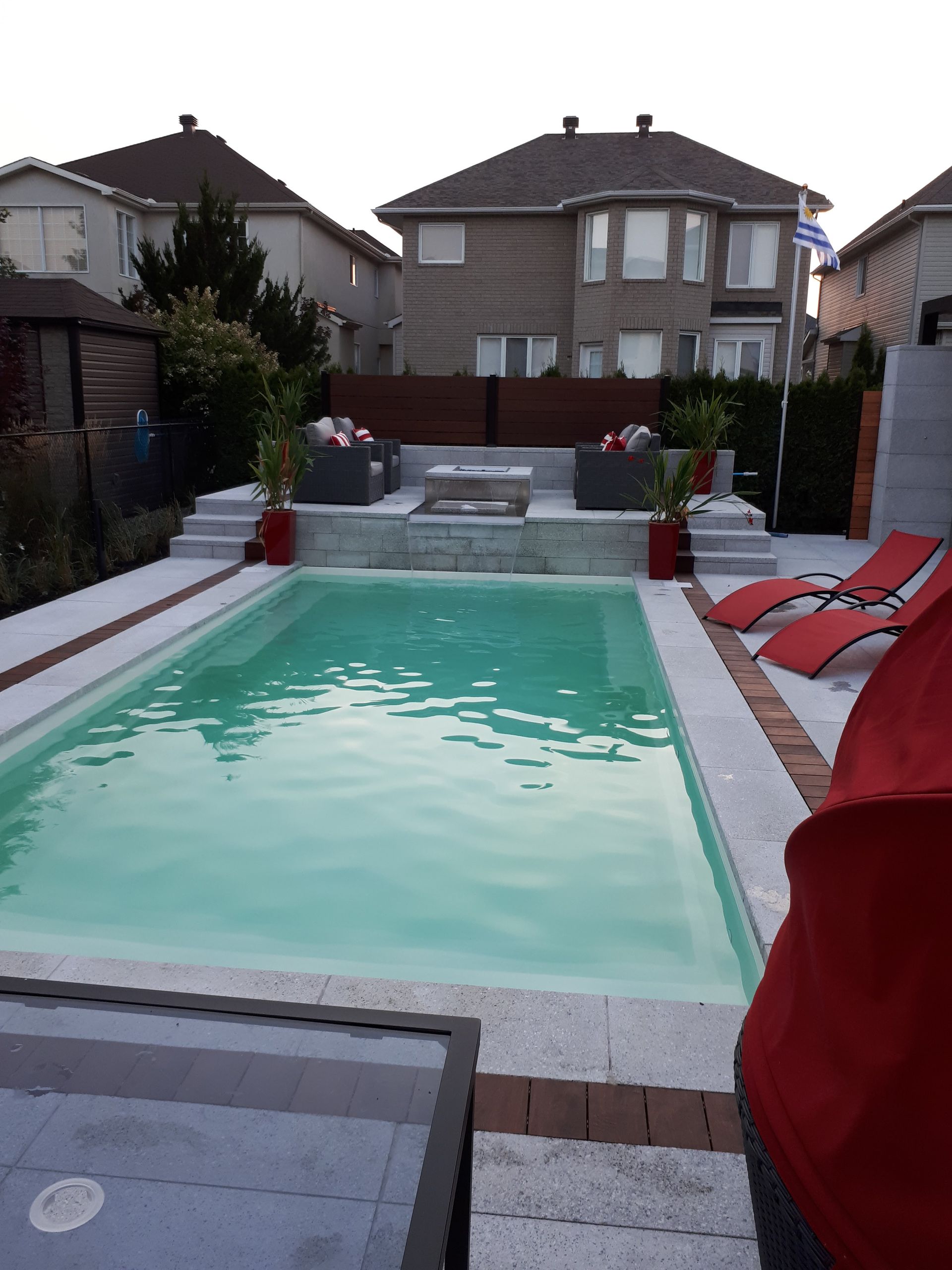 Une piscine dans le jardin avec une terrasse en pierre, un coin salon surélevé avec un foyer et des chaises longues.