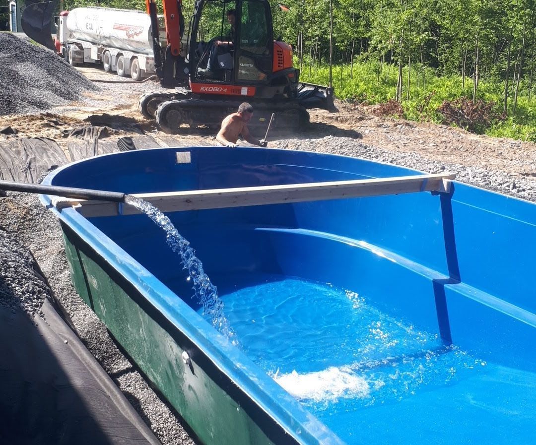 Un camion-citerne remplit une piscine en fibre de verre bleu vif, nouvellement installée sur un chantier recouvert de gravier.