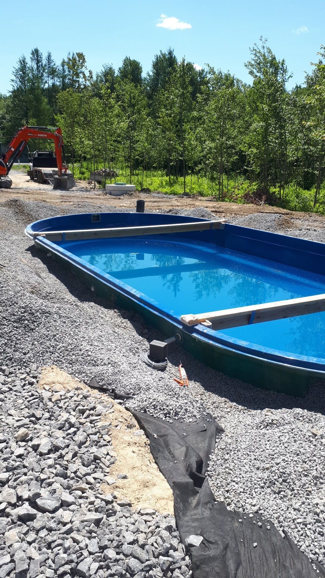Une coque de piscine en fibre de verre bleue installée sur un chantier de construction rempli de gravier, avec une excavatrice en arrière-plan.