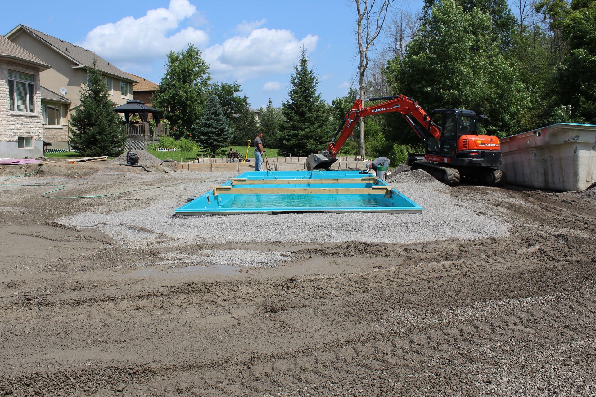 Une pelleteuse orange garée à côté d'une coque de piscine rectangulaire bleue, installée sur un chantier de construction rempli de gravier.