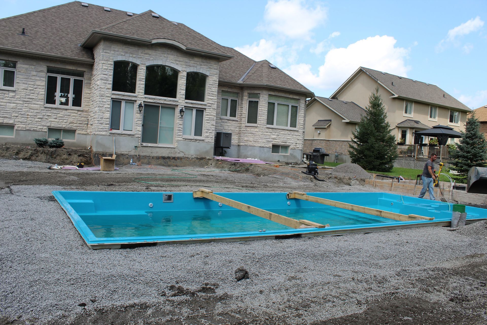Une piscine rectangulaire bleue installée dans un jardin résidentiel, entourée de gravier et de maisons voisines.