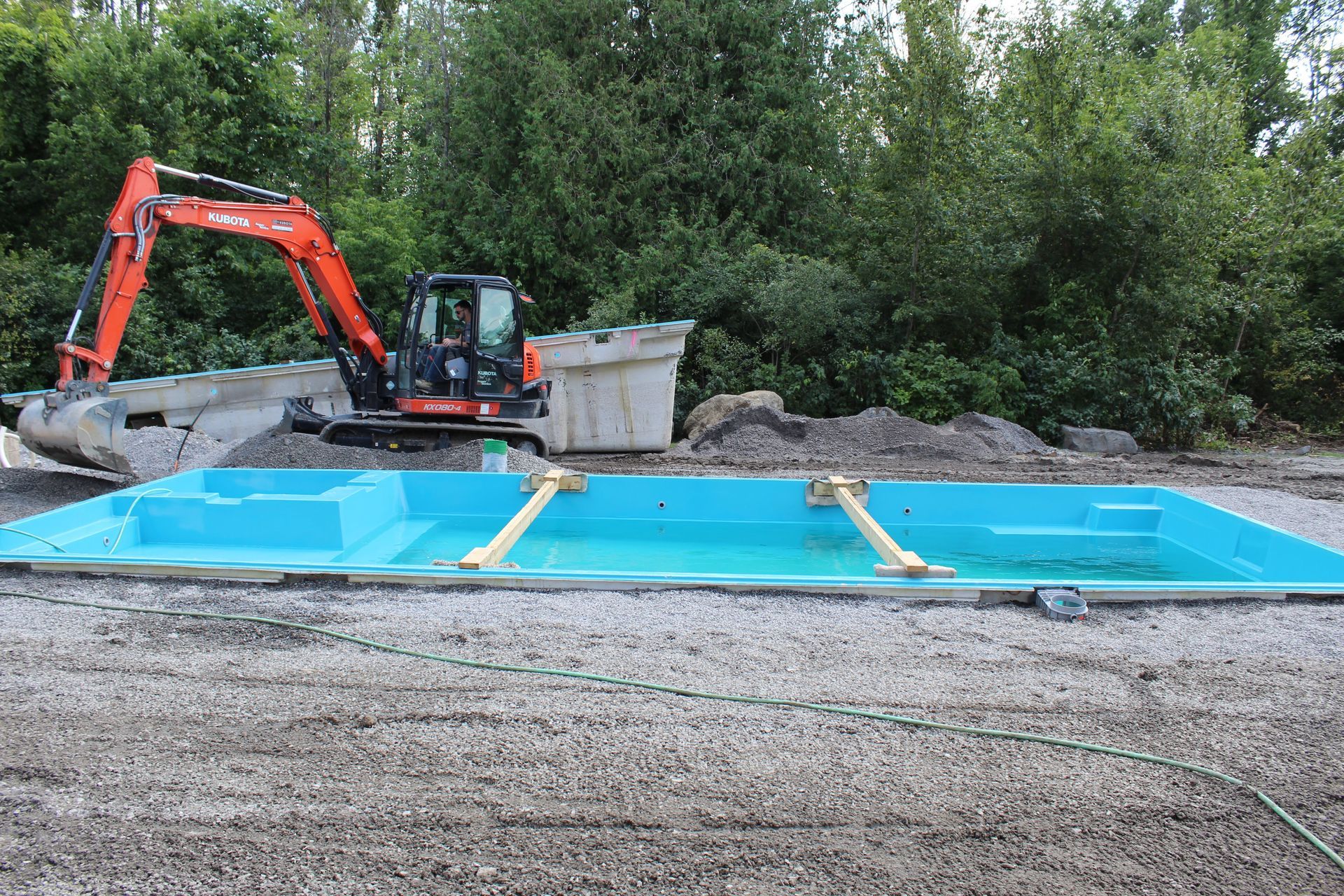 Une coque de piscine en fibre de verre bleu vif trône sur un terrain gravillonné, une pelleteuse orange étant garée à proximité.