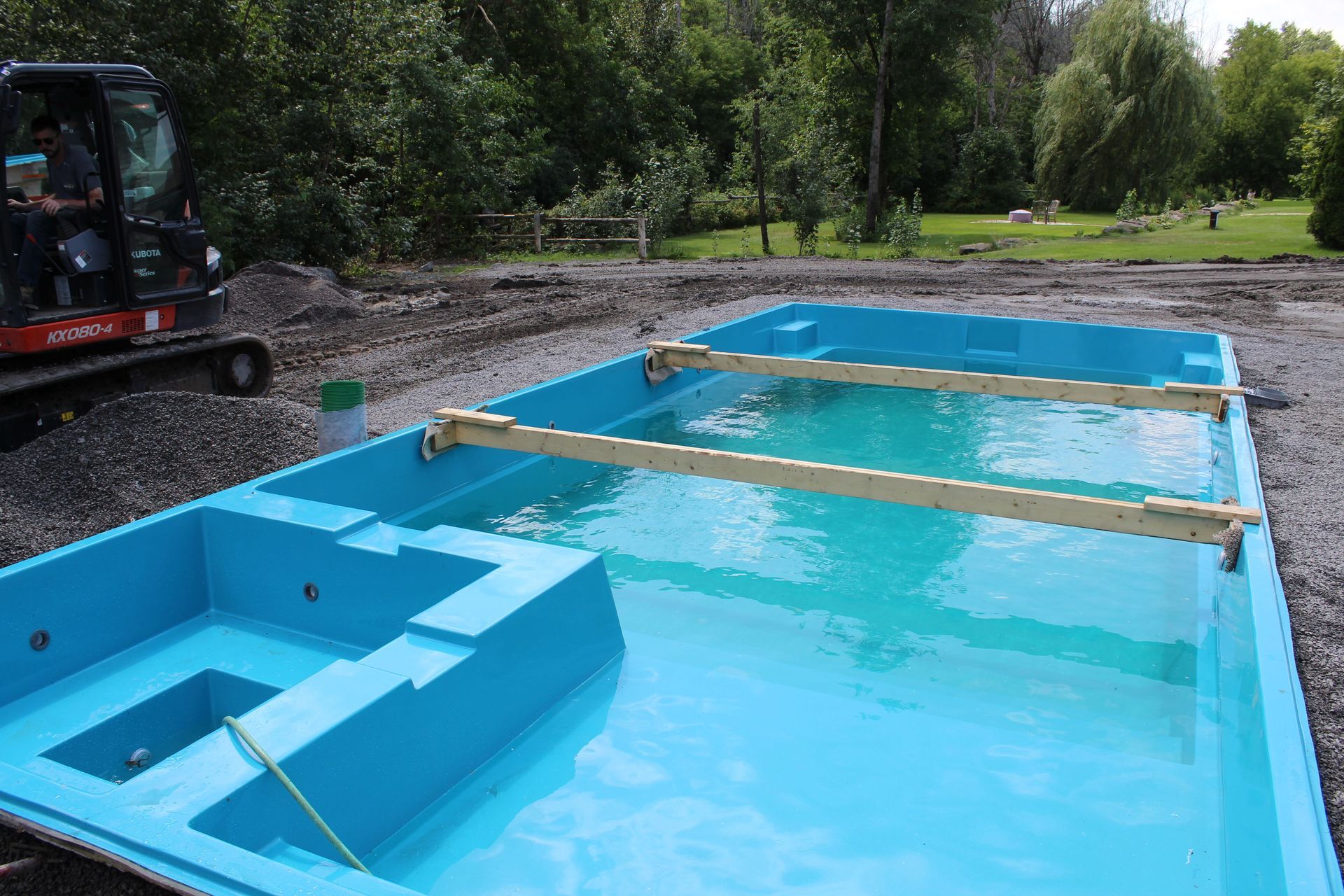 Une coque de piscine en fibre de verre bleu vif est installée sur un terrain gravillonné, soutenue par des poutres en bois sur le dessus.