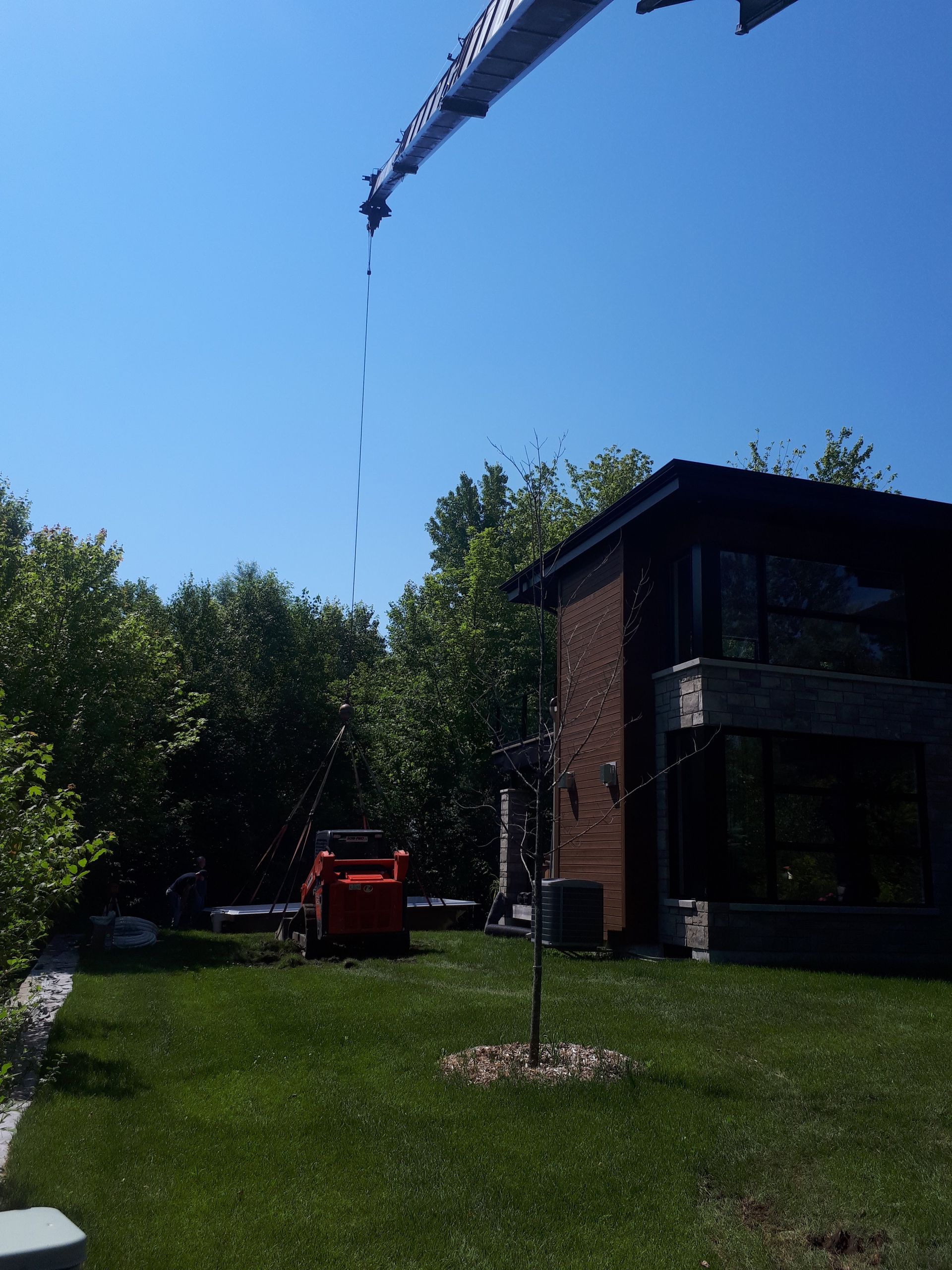 Une grue surplombe une maison et des arbres dans un jardin ensoleillé, tandis qu'une petite chargeuse compacte orange est garée sur la pelouse.