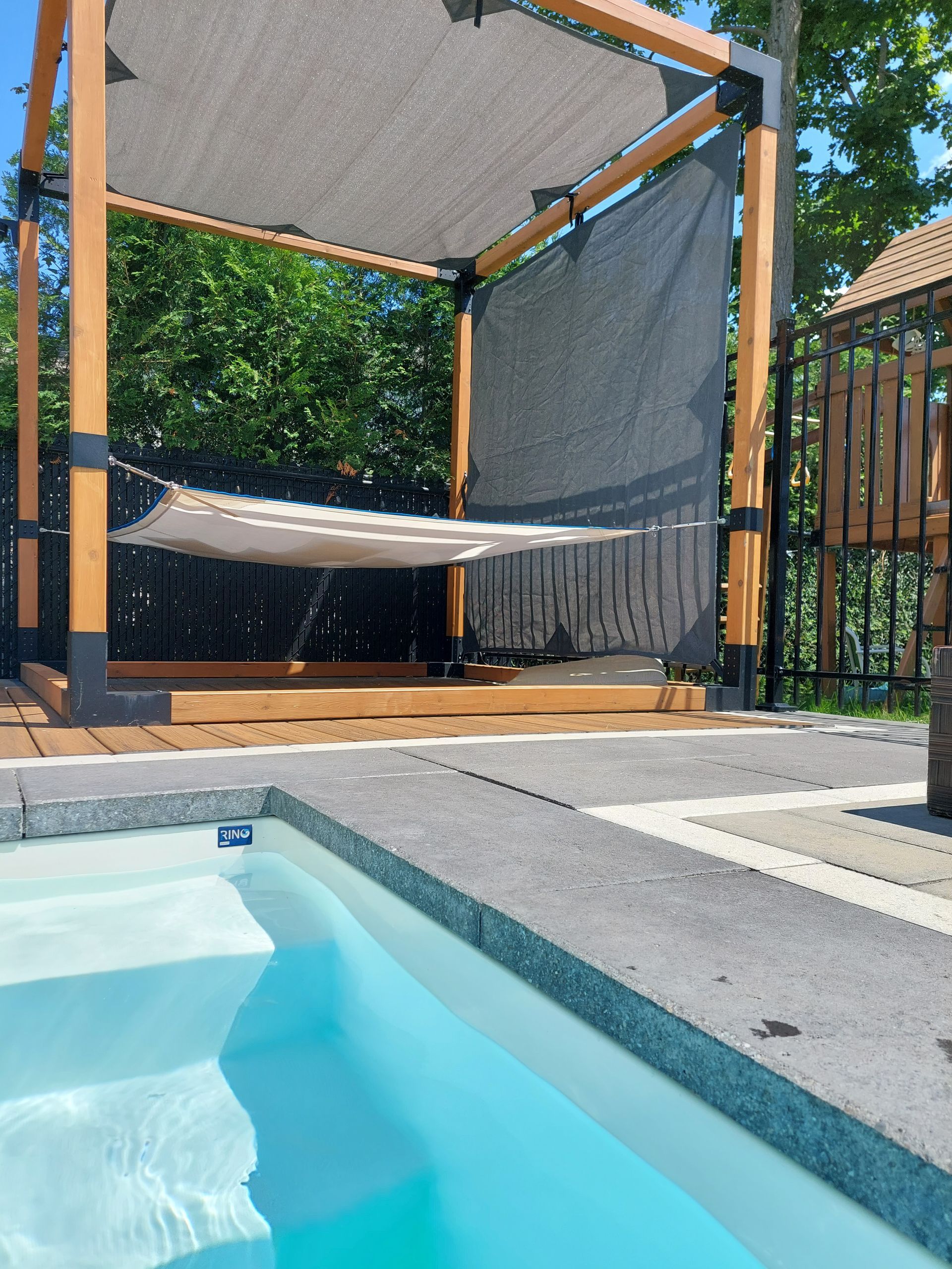 Par une journée ensoleillée, une pergola en bois avec un hamac et des toiles d'ombrage se dresse au bord d'une piscine d'un bleu éclatant.