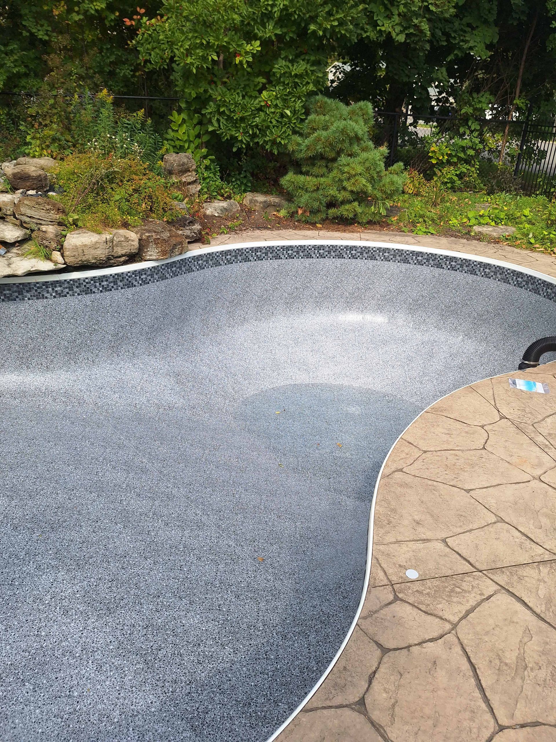 Vue en plongée d'une piscine vide, parsemée de taches grises, bordée de pavés texturés beiges et d'aménagements paysagers verdoyants.